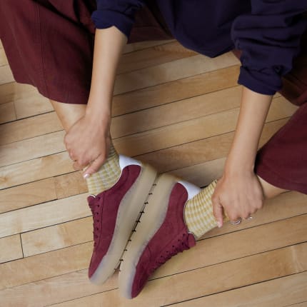 Person sitting on a wood floor wearing burgundy sneakers with a gum sole and contrasting yellow socks.